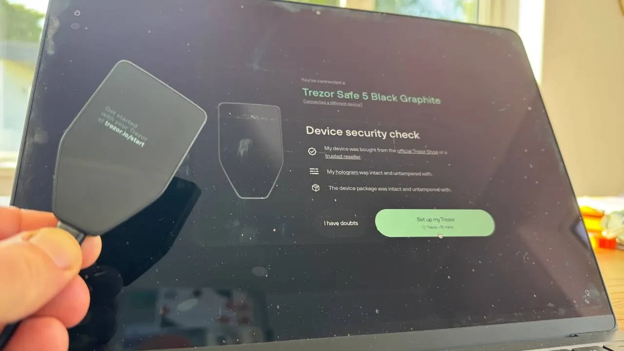 A Trezor Safe 5 held up in front of a laptop running the companion app's device security check, with checklist items confirming the hologram and packaging were intact before setup continues.