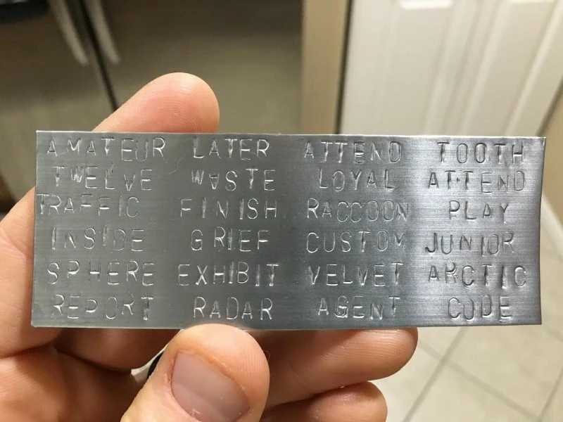 A steel seed-phrase backup plate with words stamped by hand into rows of letter tiles.